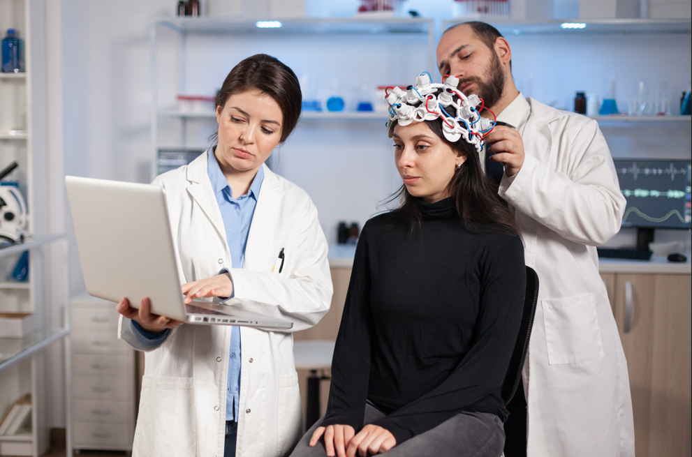 Two doctors conduct a brain study on a woman wearing a high-tech EEG headset. One doctor adjusts the device while the other reviews results on a laptop in a modern lab setting