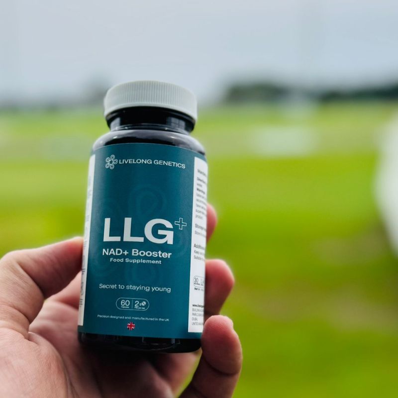 Hand holding a bottle of LLG+ NAD+ Booster supplement with a blurred green field background