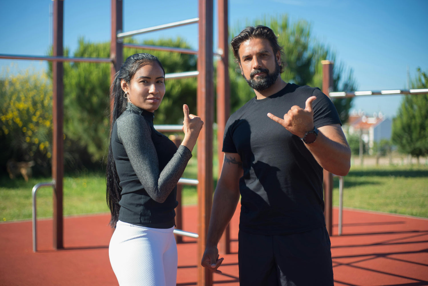 Fitness duo at outdoor workout bars — smiling and gesturing with thumbs and pinkies. Clear sky, green trees, and urban buildings frame the scene.
